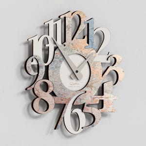 Könnte beinhalten: Eine dekorative Wanduhr mit einzigartigem Design. Die Uhr hat große, stilisierte Zahlen in Weiß und ein strukturiertes, mehrfarbiges Zifferblatt. Die Zeiger sind hellgrau, und das Zifferblatt trägt den Text "MADE IN ITALY".