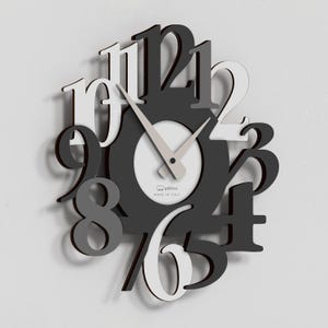 Puede incluir: Un reloj de pared moderno con un diseño único. El reloj presenta números grandes y estilizados en tonos de blanco, gris y negro. La esfera del reloj es blanca con manecillas grises claras. El texto "MADE IN ITALY" es visible.