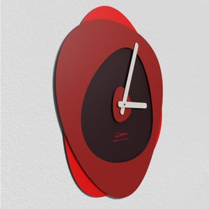 Op de afbeelding: Een moderne wandklok met een uniek, abstract ontwerp. De klok heeft een gelaagde, rode ovale vorm met een donkerbruin centrum en witte wijzers. Gemaakt in Italië.