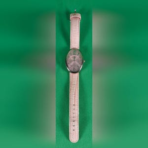 May include: An oval-shaped wristwatch featuring a silver-tone metal case and a light pink textured leather strap. The watch face has a light pink background with dark green Arabic numerals and thin silver-tone hands for hour and minute tracking.