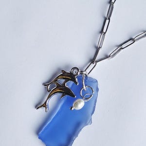 May include: A silver necklace featuring a blue glass pendant. Two dolphin charms with brown accents are attached to the pendant, along with a small pearl. The necklace has a silver chain.