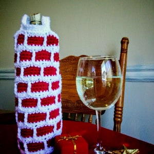 May include: A red and white crocheted wine bottle cosy, a glass of white wine, a small red gift box with a gold bow, and a gold star ornament are on a red tablecloth. A wooden chair is in the background.