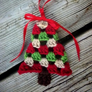 May include: A crocheted Christmas tree ornament in red, green, and cream with a brown yarn trunk. The ornament is attached to a red ribbon with a bow.