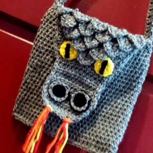 May include: A gray crocheted dragon-shaped bag with yellow eyes and black nostrils. The bag has a red and yellow fringe hanging from the bottom.