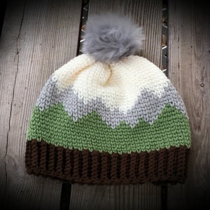 Op de afbeelding: Een gebreide wintermuts met een bruin, groen, grijs en wit kleurpatroon. De muts heeft een pluche grijze pompon bovenop.
