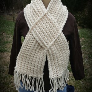 May include: A white crocheted scarf with fringe on the ends. The scarf is folded in a criss-cross pattern and is being worn by a person.