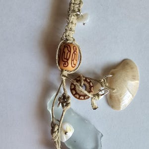 May include: A handcrafted macrame pendant featuring natural elements. It includes a variety of beads, a piece of sea glass, and a seashell. The pendant is suspended from a beige rope, showcasing a bohemian aesthetic.