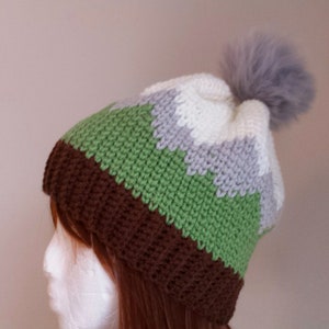 Op de afbeelding: Een gebreide beanie met bruine, groene, grijze en witte strepen en een pluche grijze pompon bovenop.