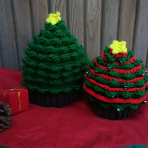 Peut inclure: Deux décorations de sapin de Noël crochetées. L'une est un grand sapin vert avec une étoile jaune au sommet, et l'autre est un sapin plus court avec des rayures rouges et vertes, orné de petites perles. Une petite boîte cadeau rouge et une pomme de pin sont également présentes.