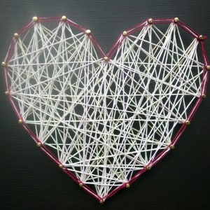 May include: A heart-shaped string art piece with white and light pink string wrapped around gold tacks on a dark background. The tacks are arranged in the shape of a heart, and the string is wrapped around them in a random pattern.