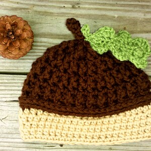 May include: A brown and beige crocheted acorn hat with a green crocheted leaf on top. The hat is sitting on a wooden surface.