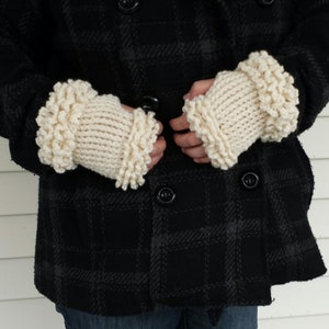May include: A pair of white knitted fingerless gloves with a ruffled edge. The gloves are worn by a person wearing a black and gray plaid coat.