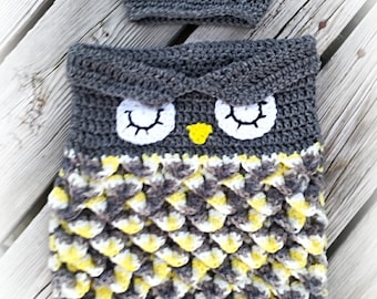 May include: A gray crocheted owl baby sleeping bag with yellow and white accents. The sleeping bag has a matching gray crocheted hat with yellow and white ties.