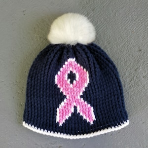 Puede incluir: Un gorro de punto azul marino con un pompón blanco en la parte superior. El gorro tiene un diseño de cinta rosa en la parte delantera.