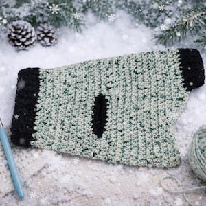 May include: A handmade crochet dog sweater in shades of green and white with black trim. The sweater has a hole for the leash and is surrounded by yarn, a crochet hook, pine cones, and faux snow. The sweater is a cozy winter accessory.