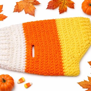 Könnte beinhalten: Ein gehäkelter Hundepullover in den Farben von Candy Corn: Weiß, Orange und Gelb. Der Pullover hat ein Loch für die Leine. Das Bild ist umgeben von orangefarbenen Kürbissen, weißen Kürbissen, Candy Corn und Herbstblättern.
