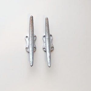 May include: Two silver metal cleat hooks, each with four holes and a pointed end. The hooks are mounted on a white background.