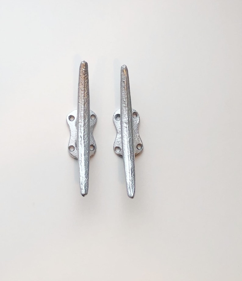 May include: Two silver metal cleat hooks, each with four holes and a pointed end. The hooks are mounted on a white background.