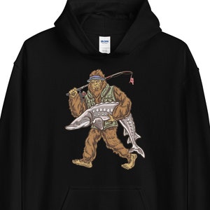 Puede incluir: Sudadera con capucha negra con un estampado gr&aacute;fico de un Bigfoot sosteniendo un pez grande. El Bigfoot lleva un chaleco verde y un sombrero.