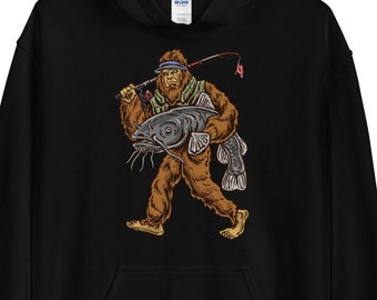 Puede incluir: Sudadera con capucha negra con un gráfico de un Bigfoot sosteniendo un pez gato. El Bigfoot lleva un sombrero y sostiene una caña de pescar.