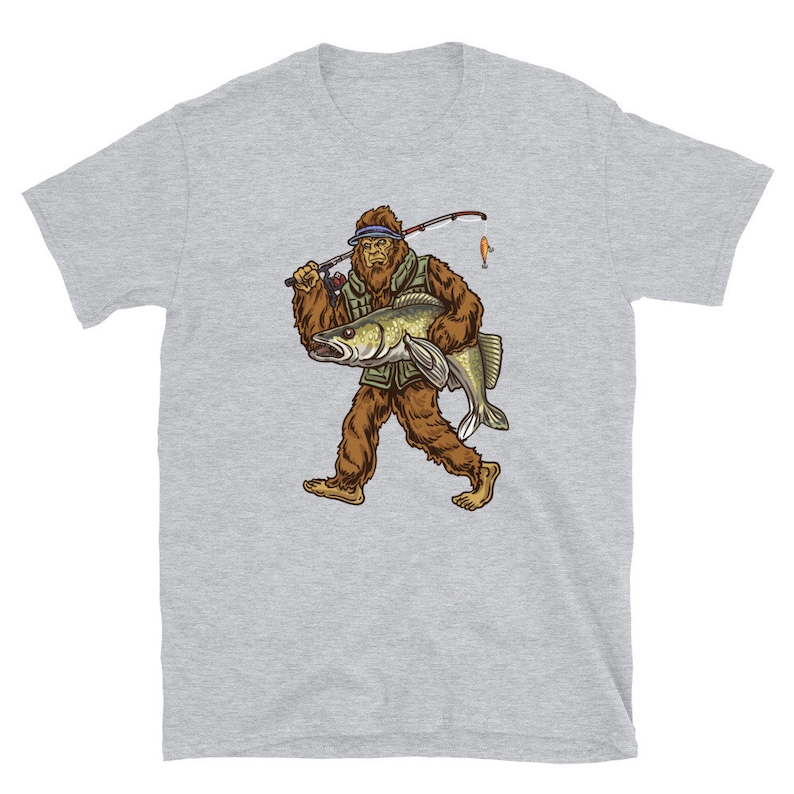 Op de afbeelding: Een grijs T-shirt met een afbeelding van een Bigfoot die een vis en een hengel vasthoudt. De Bigfoot draagt een groene vest en een blauwe hoed.