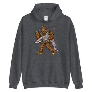 Puede incluir: Una sudadera con capucha gris oscuro con un estampado gr&aacute;fico de un Bigfoot sosteniendo un pez grande. El Bigfoot lleva un chaleco verde y un pa&ntilde;uelo. El pez es gris y tiene un hocico largo.