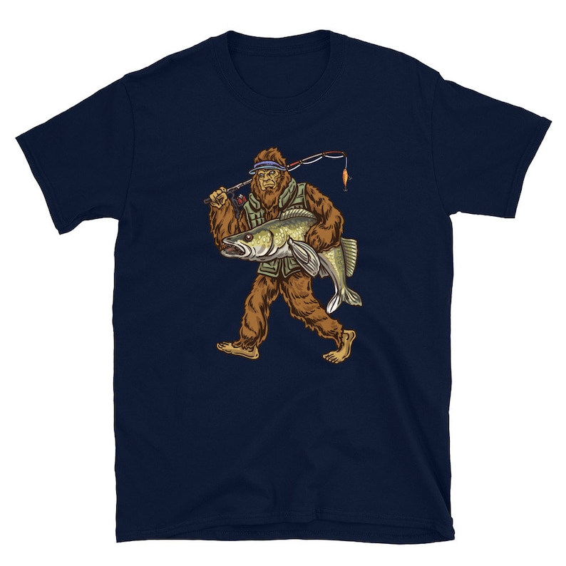 Op de afbeelding: Een marineblauw t-shirt met een afbeelding van Bigfoot die een vis en een hengel vasthoudt. Bigfoot draagt een vissersvest en een hoed.