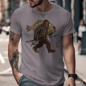 Könnte beinhalten: Graues T-Shirt mit einer Illustration eines Bigfoot, der einen großen Fisch und eine Angelrute trägt. Das Design verwendet Braun-, Grün- und Beigetöne. Eine kleine Garnele ist ebenfalls abgebildet. Das T-Shirt ist ein Freizeitkleidungsstück.
