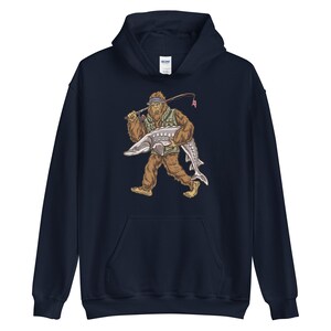 Puede incluir: Una sudadera con capucha azul marino con un gr&aacute;fico de un Bigfoot sosteniendo un pez grande. El Bigfoot lleva un chaleco verde y un sombrero.