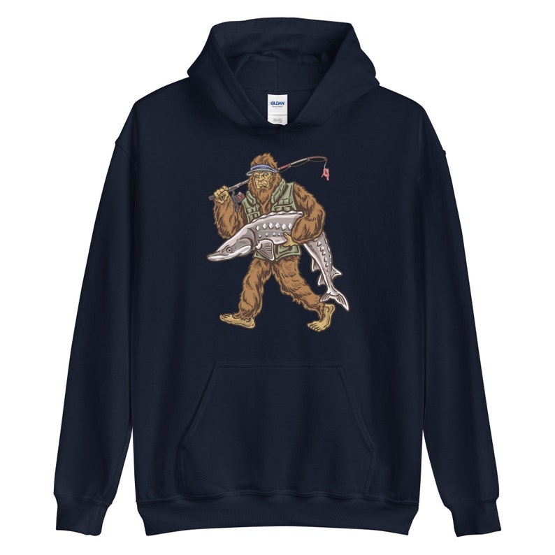 Puede incluir: Una sudadera con capucha azul marino con un gr&aacute;fico de un Bigfoot sosteniendo un pez grande. El Bigfoot lleva un chaleco verde y un sombrero.