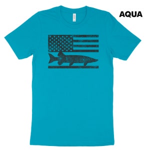 Puede incluir: Una camiseta azul turquesa con un gr&aacute;fico en blanco y negro desgastado de un pez y una bandera estadounidense. El texto "AQUA" est&aacute; en la esquina superior derecha de la imagen.