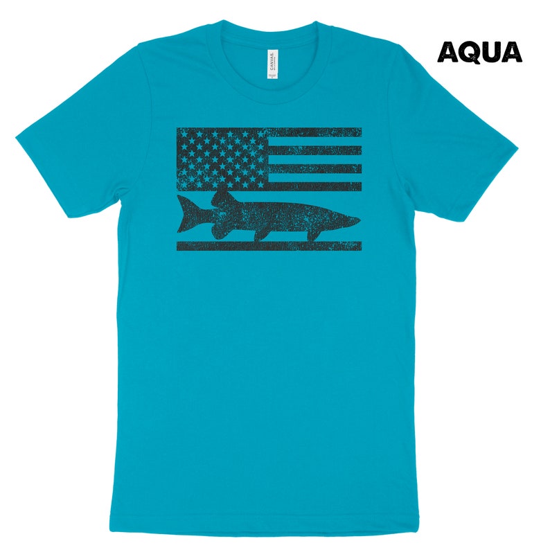 Puede incluir: Una camiseta azul turquesa con un gr&aacute;fico en blanco y negro desgastado de un pez y una bandera estadounidense. El texto "AQUA" est&aacute; en la esquina superior derecha de la imagen.