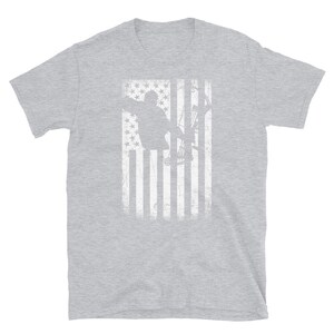 May include: A light gray t-shirt featuring a distressed white graphic of an archer with a bow and arrow, set against a stylized American flag background. The design has a vintage, worn look.