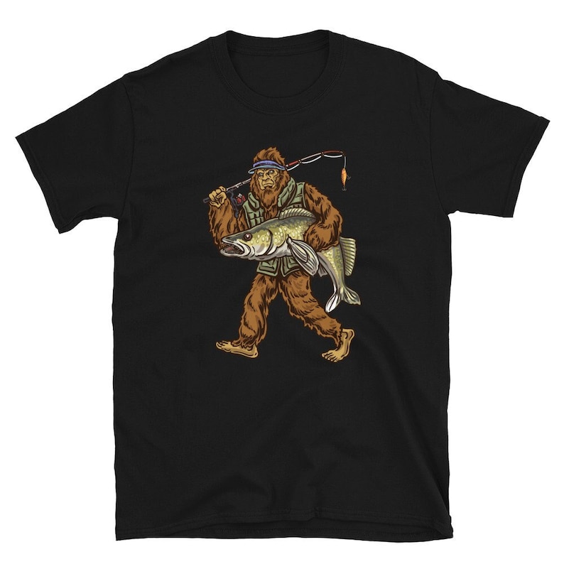 Op de afbeelding: Zwart T-shirt met een afbeelding van een Bigfoot die een vis vasthoudt. De Bigfoot draagt ​​een hoed en houdt een hengel vast.