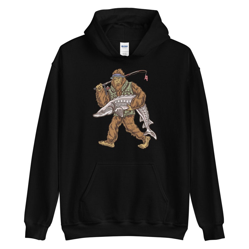 Puede incluir: Sudadera con capucha negra con un estampado gr&aacute;fico de un Bigfoot sosteniendo un pez grande. El Bigfoot lleva un chaleco verde y un sombrero.