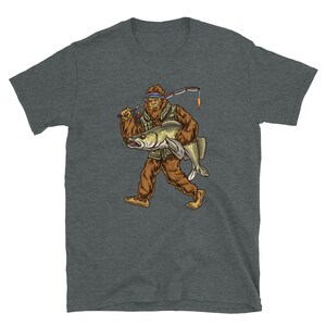 Op de afbeelding: Een donkergrijs T-shirt met een afbeelding van Bigfoot die een vis en een hengel vasthoudt. Bigfoot draagt een groene vest en een bruine hoed.