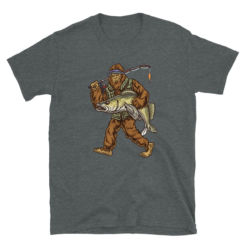Op de afbeelding: Een donkergrijs T-shirt met een afbeelding van Bigfoot die een vis en een hengel vasthoudt. Bigfoot draagt een groene vest en een bruine hoed.