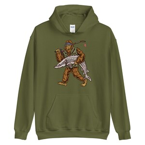 Puede incluir: Una sudadera con capucha verde con un estampado gr&aacute;fico de un Bigfoot de dibujos animados sosteniendo un pez grande. El Bigfoot lleva un chaleco de pesca y un sombrero. El pez es un esturi&oacute;n.