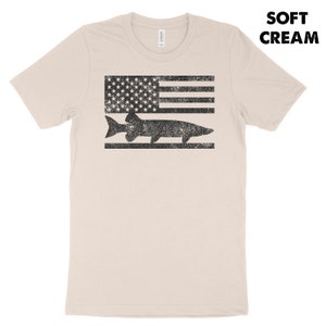 Puede incluir: Una camiseta de color crema suave con un gr&aacute;fico negro y blanco desgastado de un pez nadando sobre una bandera estadounidense estilizada.