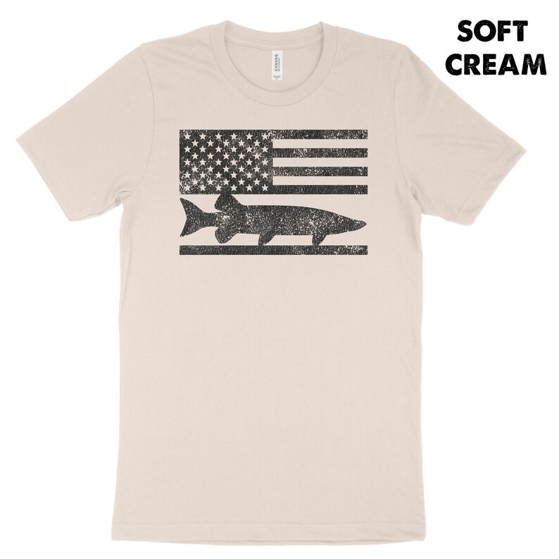 Puede incluir: Una camiseta de color crema suave con un gr&aacute;fico negro y blanco desgastado de un pez nadando sobre una bandera estadounidense estilizada.