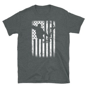 May include: Dark gray t-shirt featuring a distressed American flag design. The flag graphic includes a silhouette of a person aiming a bow and arrow. The design is in white, creating a patriotic and sporty look.