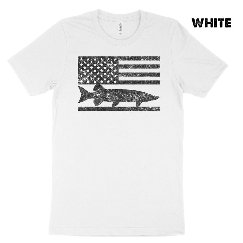 Puede incluir: Camiseta blanca con un dise&ntilde;o de bandera estadounidense desgastado que presenta una silueta negra de un pez. El texto "WHITE" est&aacute; en la esquina superior derecha de la imagen.