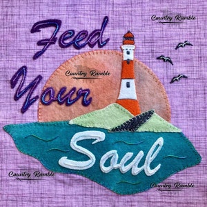 May include: A purple fabric background with a sunset and a lighthouse. The text "Feed Your Soul" is stitched in purple and white thread. The text "Country Ramble" is stitched in white thread.