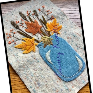 May include: A blue mason jar filled with a bouquet of autumn leaves and branches. The leaves are orange, yellow, and brown. The background is a white speckled fabric. The text "Country Knick Knacks" is written in blue on the fabric.