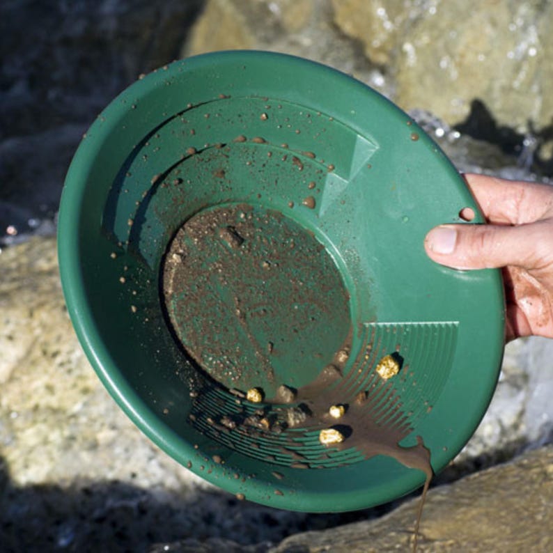 Puede incluir: Una batea de pl&aacute;stico verde llena de tierra y peque&ntilde;as pepitas de oro. La batea es sostenida por una mano, con agua y rocas en el fondo. Se utiliza para la prospecci&oacute;n de oro.