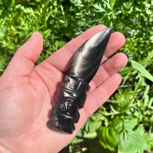 May include: A black obsidian dagger with gold flecks is held in a hand. The dagger has a detailed handle and a pointed blade. The background is blurred green foliage.