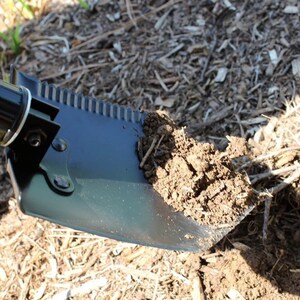 May include: A black folding shovel with a serrated edge is being used to dig into the ground. The shovel has a black handle and a metal blade.