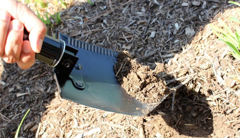 May include: A black folding shovel with a serrated edge is being used to dig into the ground. The shovel has a black handle and a metal blade.