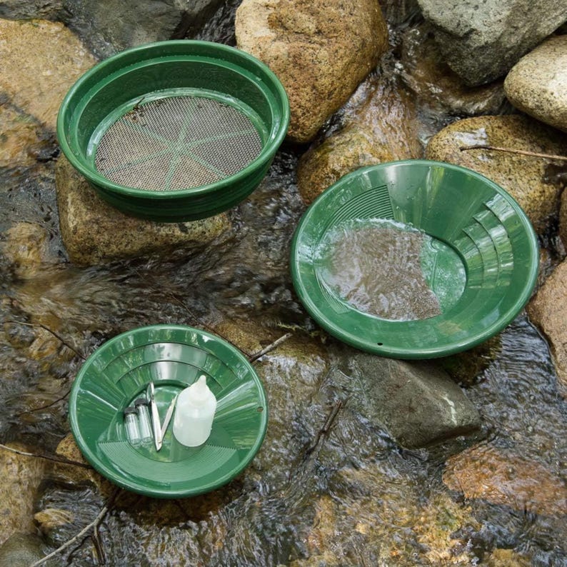 Puede incluir: Un conjunto de herramientas verdes para la b&uacute;squeda de oro, que incluye un tamiz, dos bandejas, una botella peque&ntilde;a y unas pinzas. Las herramientas est&aacute;n dispuestas sobre rocas en un arroyo, sugiriendo uso al aire libre.