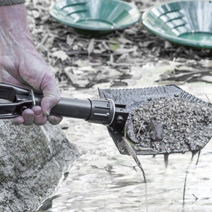 May include: A black folding shovel with a metal handle is being used to scoop up gravel from a stream. The shovel is filled with gravel and water is dripping from the edge. Two green metal pans are in the background.